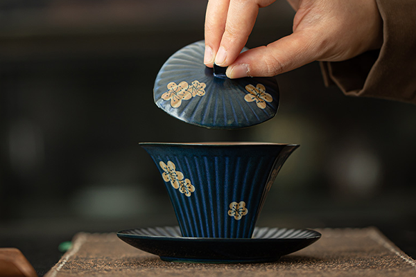 濃い青色の梅の花模様入り蓋碗の蓋を、人が操作しているところ。