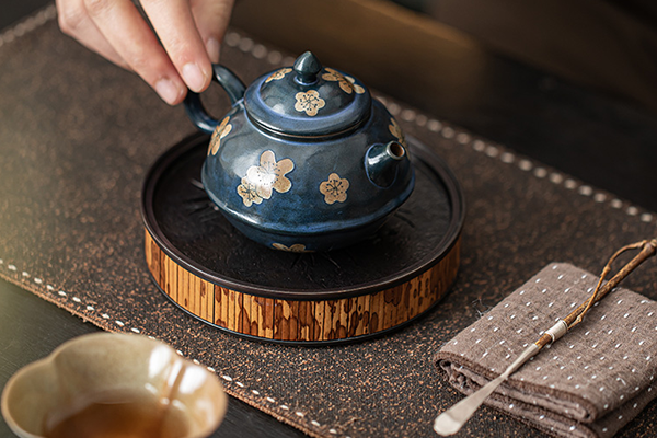 茶盤の上に置かれた、粉引青釉の氷梅文様入り茶壺に手が添えられている。茶道具や茶托も一緒に写っており、茶席の風景が感じられる。
