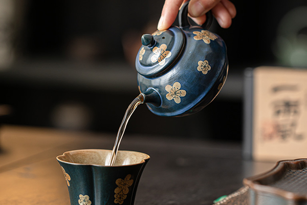 茶盤の上に置かれた、粉引青釉の氷梅文様入り茶壺に手が添えられている。茶道具や茶托も一緒に写っており、茶席の風景が感じられる。