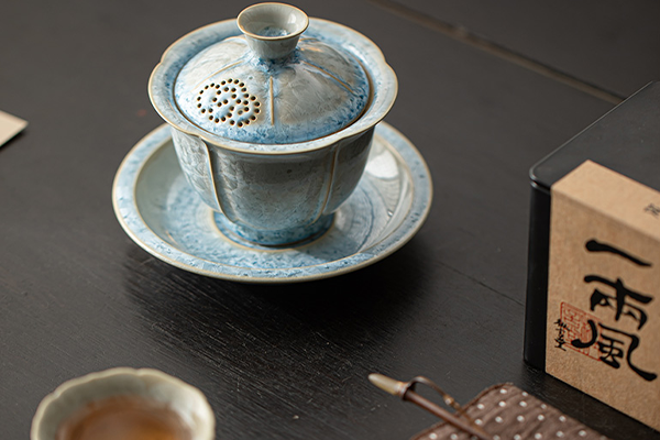 黒いテーブルの上に置かれた、青い結晶釉の蓋碗セットと茶葉の箱。蓋には透かし彫りの穴があり、趣のある雰囲気。
