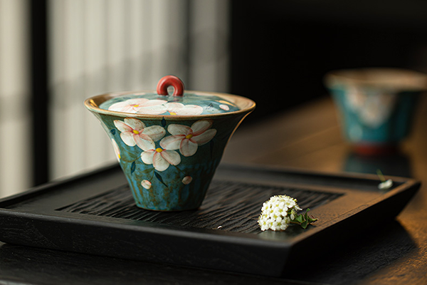 黒い茶盤の上に置かれた、青藍色の「手描き繁花」模様の太湖石蓋碗。手前には小さな白い花が添えられている。