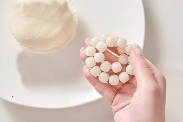 白いプレートの上に置かれた白いデザートの横で、手が明るいクリーム色の菩提根ビーズで作られたブレスレットを指で回しているクローズアップ。