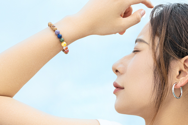 青い空を背景に、目を閉じて顔を上げ、片手を額にかざしている女性の横顔。左手首には、茶色の木製ビーズと、紫、青、緑、黄、オレンジ、赤の七色の石のビーズが連なったブレスレットを着用している。