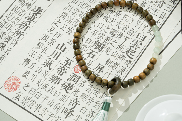 緑檀のビーズと薄緑色の模様のある石のビーズで作られ、暗い色の木のリングと長い白い房がアクセントになったビーズのブレスレットが、中国語の伝統的な文字が書かれた開いた本または巻物の上に置かれている。右下隅には小さな白い皿が一部見えている。