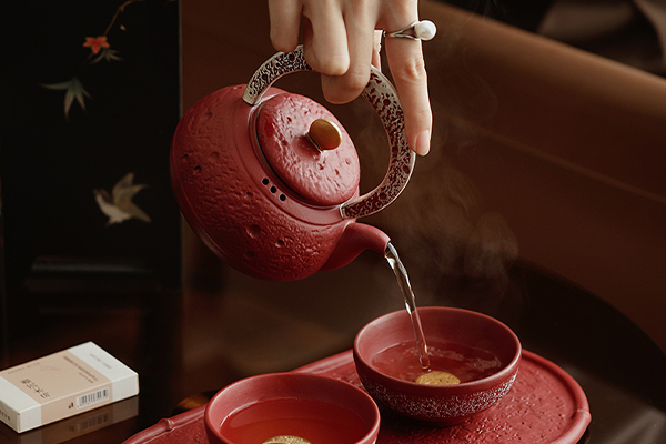 提梁式急須から赤い茶碗にお茶が注がれている様子。茶碗の中には金色の丸い模様が見える。湯気が立ち上り、温かいお茶の香りが漂うような雰囲気。