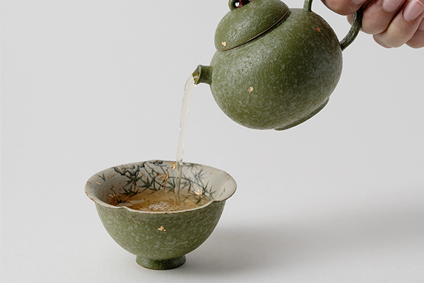 抹茶色の急須から、金彩散らし湯呑へお茶が注がれている様子。湯呑の内側には竹の葉のような絵柄が描かれ、金色の箔が散らされている。