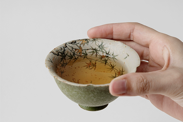 手に持たれた橘緑釉・金彩散らし湯呑。中にはお茶が注がれており、湯呑の内側に描かれた竹の葉と金箔が、お茶の琥珀色と調和している。