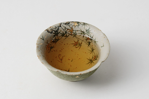 白い背景に置かれた、お茶が注がれた橘緑釉・金彩散らし湯呑。お茶の表面には細かな茶葉が見え、湯呑の内側の竹の葉と金箔の模様が美しい。