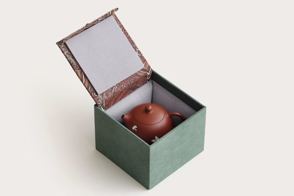 蓋が開いた状態の、緑色の箱に収められた銀継ぎの朱泥梨形急須。箱の内側には模様があり、急須は保護材に包まれて大切に収納されている様子。急須には白い花模様が描かれている。