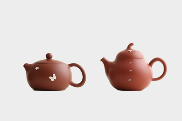 白い背景に並んだ、二つのデザインの茶色の丸い急須。一つは花と蝶の飾りが施され、もう一つは縦の線と花のような飾りがついている。