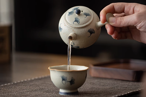 青花・火石紅 手描き銀杏文様の急須からお茶が茶碗に注がれている様子。湯気が立ち、温かいお茶の準備が感じられる。急須と茶碗には青いイチョウの葉の模様が描かれている。