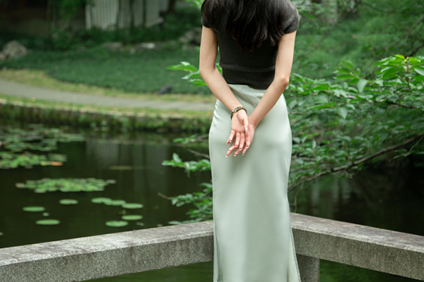 緑の水辺のほとりで、黒いトップスと淡い緑色のスカートを着た人物が、両手を後ろで組んで立っている。手首には、緑色のビーズブレスレットが見える。