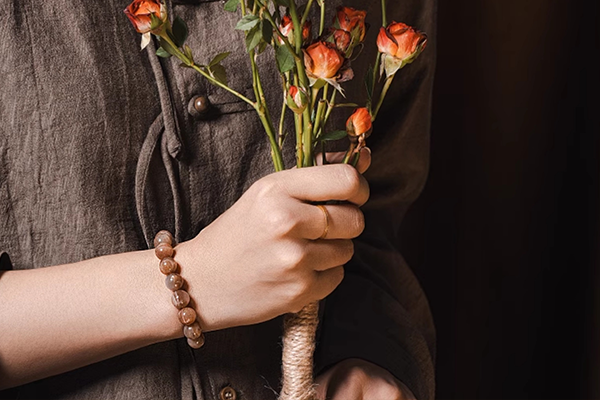 茶色い服を着た人物が、束ねられたドライフラワーのバラの花束を手に持っている。手首には茶色とオレンジ色のビーズブレスレットが着けられており、ビーズは光沢を放っている。