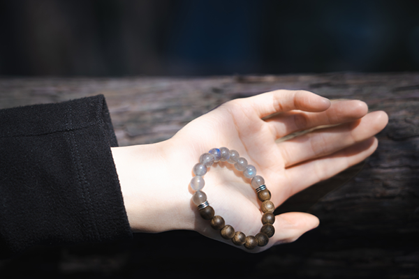 黒い服を着た人物の手のひらに、青いムーンストーンと茶色い木製ビーズでできたブレスレットが乗っている。背景には暗い木製の表面が写っている。