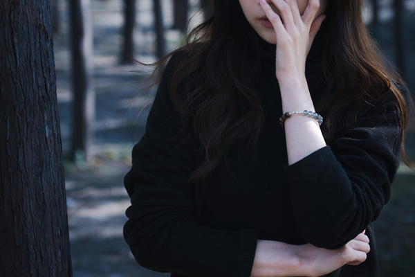 暗い森のような場所で、黒い服を着た人物が片手で顔を覆っている。手首には、青いムーンストーンと木製ビーズでできたブレスレットが着けられている。